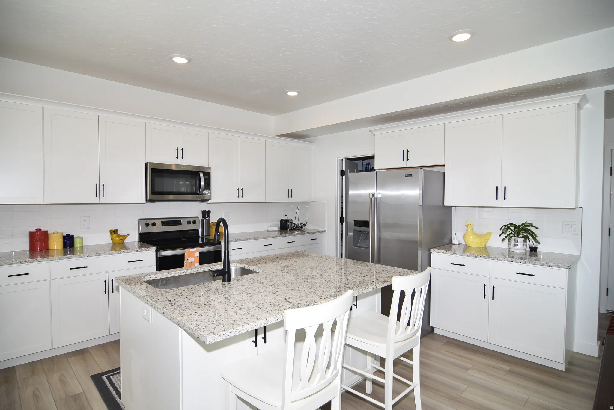 Contemporary kitchen featuring an island with a stainless steel sink and matte black faucet, white shaker cabinetry, speckled countertops, stainless steel appliances, and wood-finish flooring