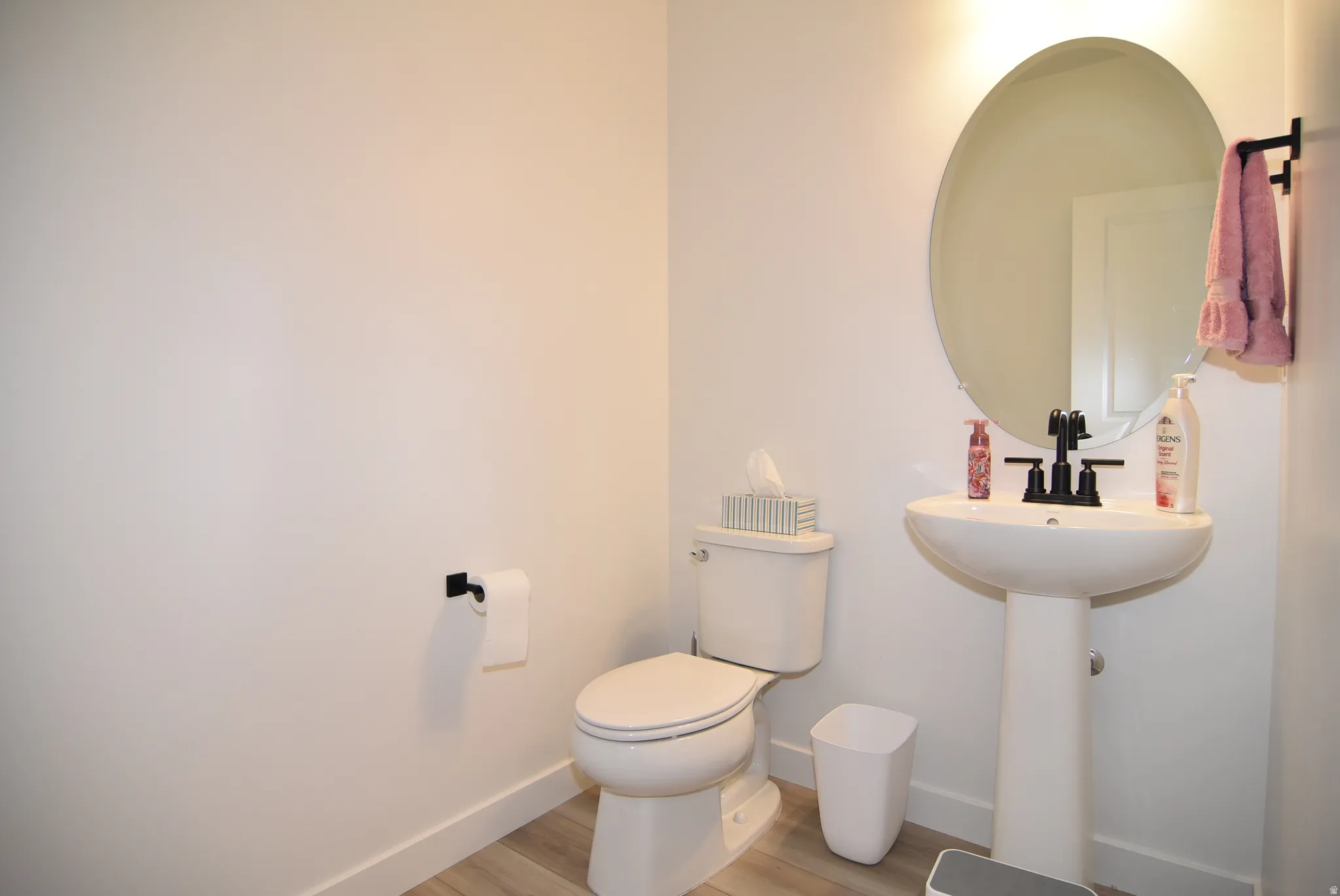Guest 1/2 Bathroom featuring a pedestal sink with a circular mirror, matte black faucet, and wood-finish flooring