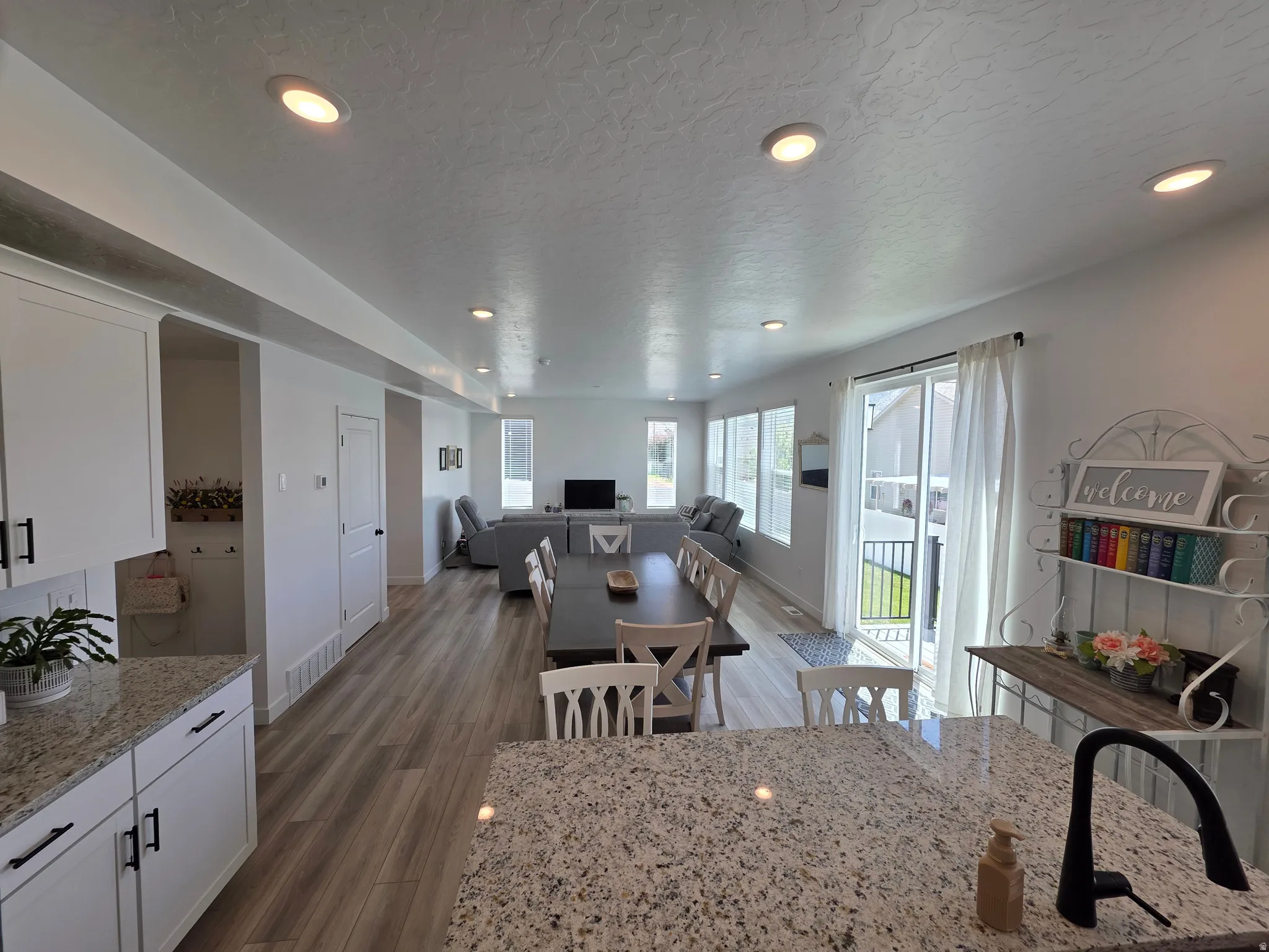 Spacious open-concept living area featuring wood-finish flooring, recessed lighting, a sliding glass door to an exterior balcony, light-toned walls, and a kitchen island with a speckled countertop and a black gooseneck faucet