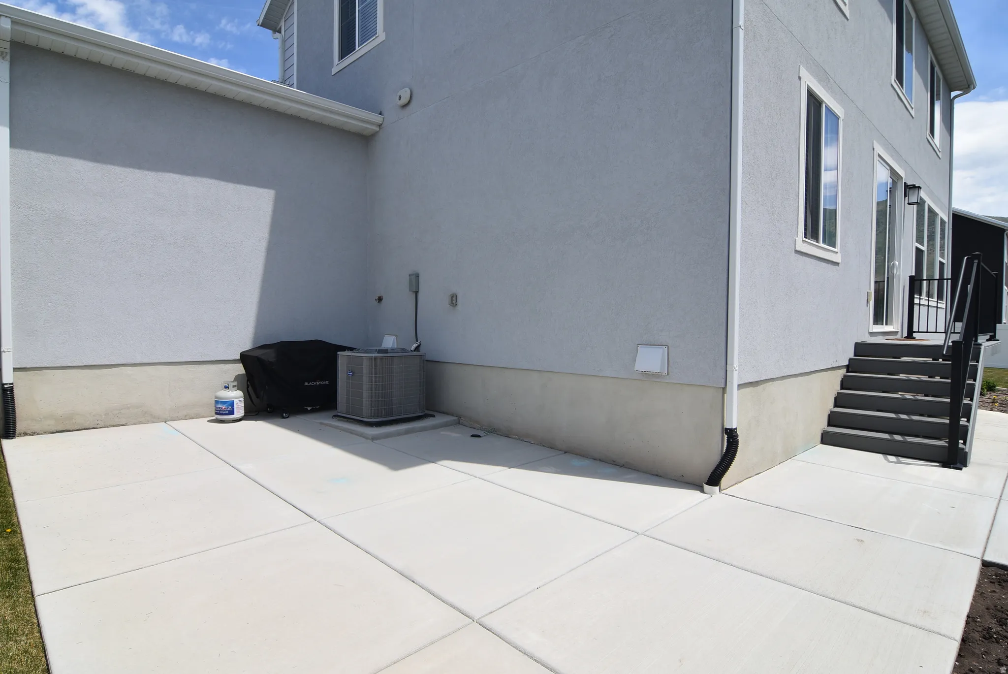 Patio area with concrete pavers, light gray stucco siding, white trim windows, and black exterior stairs with a metal railing