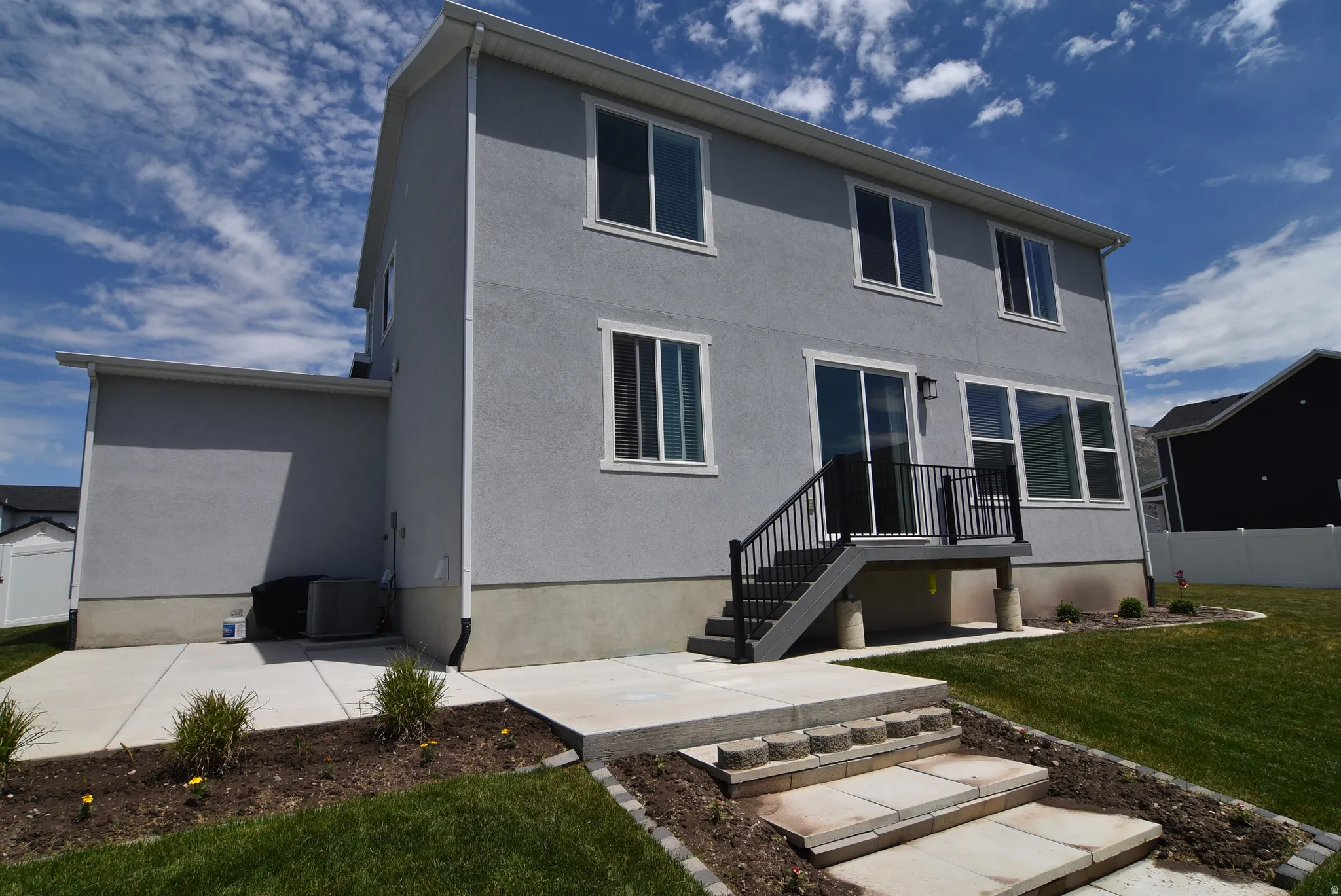 Rear exterior featuring a sliding glass door, multiple windows with white trim, a ground-level concrete patio, and an elevated deck with black railings and stairs