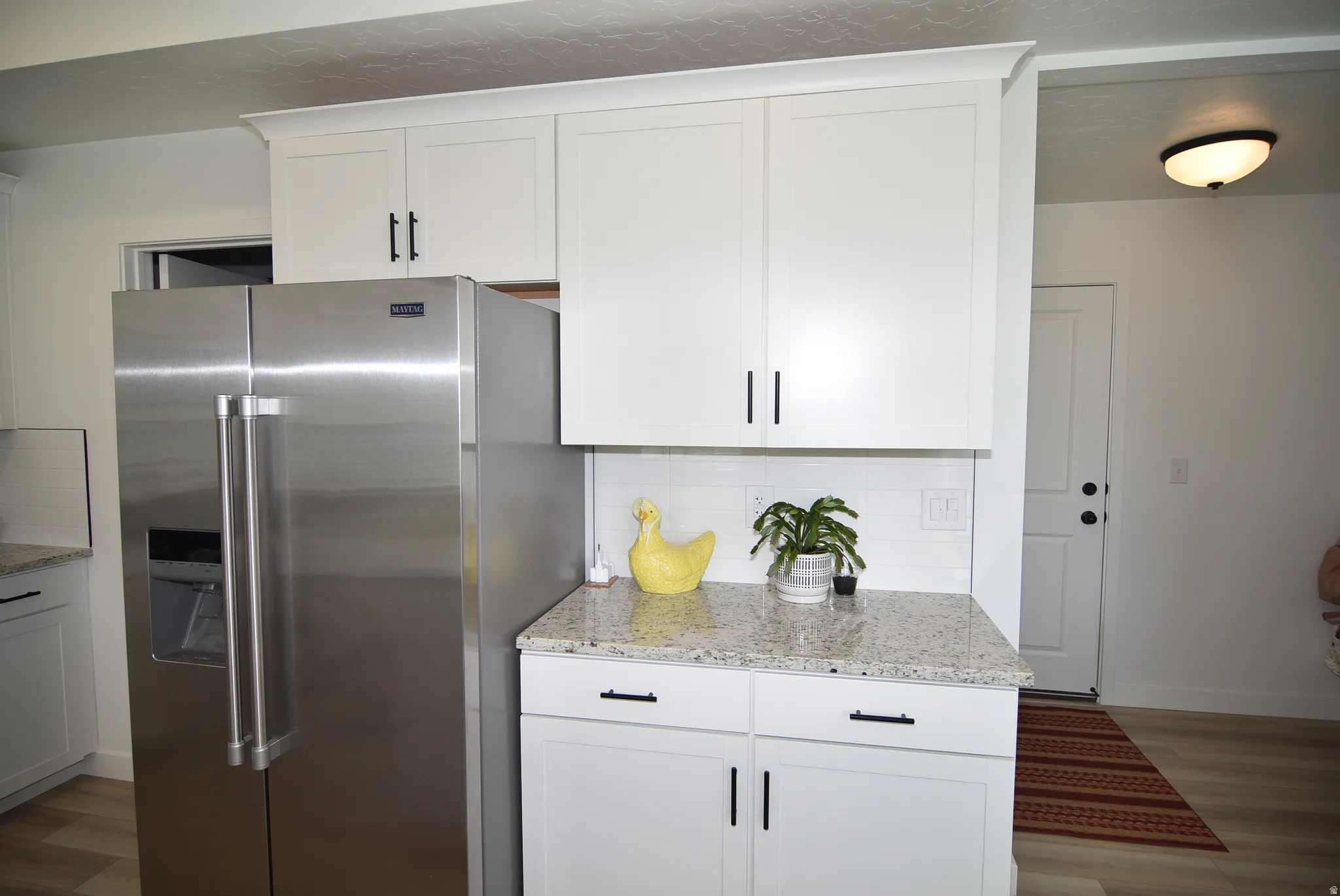 Stainless steel side-by-side refrigerator, white shaker cabinetry with black hardware, light-toned stone-finish countertop, and white subway tile backsplash