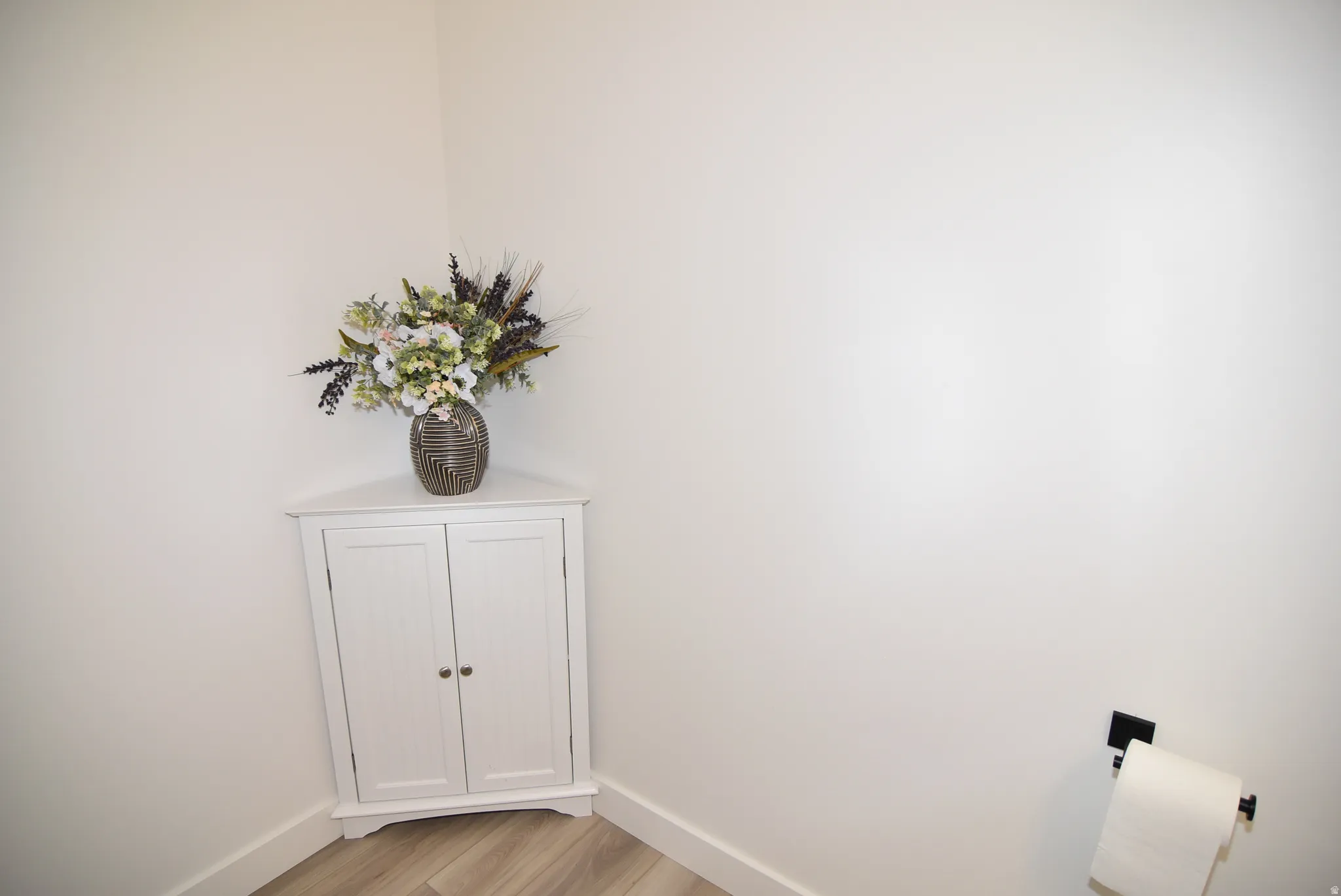 White corner cabinet with paneled doors, light wood-finish flooring, and white baseboards