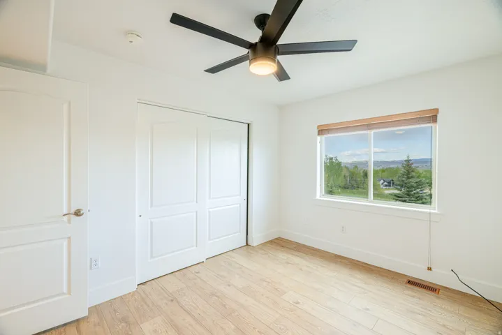 Bright interior space featuring a window with a distant landscape, wood-finish flooring, a contemporary ceiling fan with integrated lighting, a solid-core door with brushed nickel hardware, and bi-fold closet doors