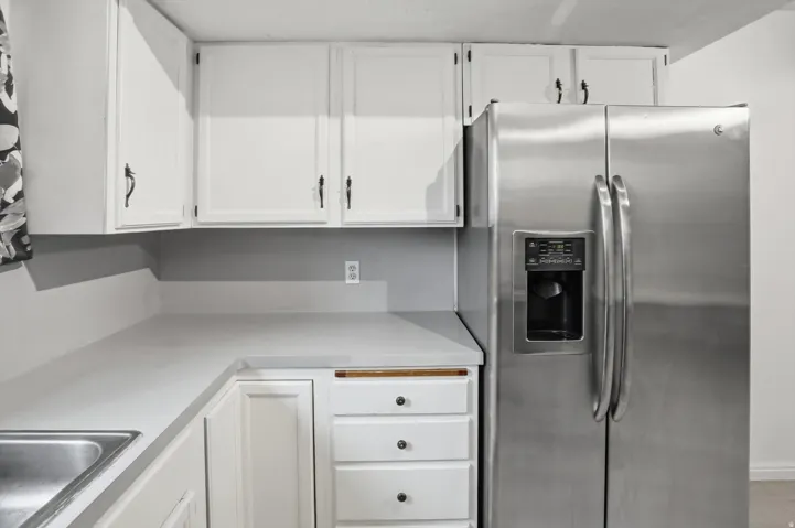 White cabinetry with black hardware, light-toned countertops, stainless steel side-by-side refrigerator with exterior dispenser, integrated stainless steel sink, and drawer storage