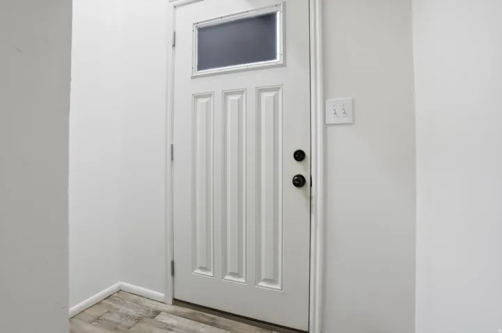 White paneled door with an upper window insert, featuring black hardware and wood-finish flooring