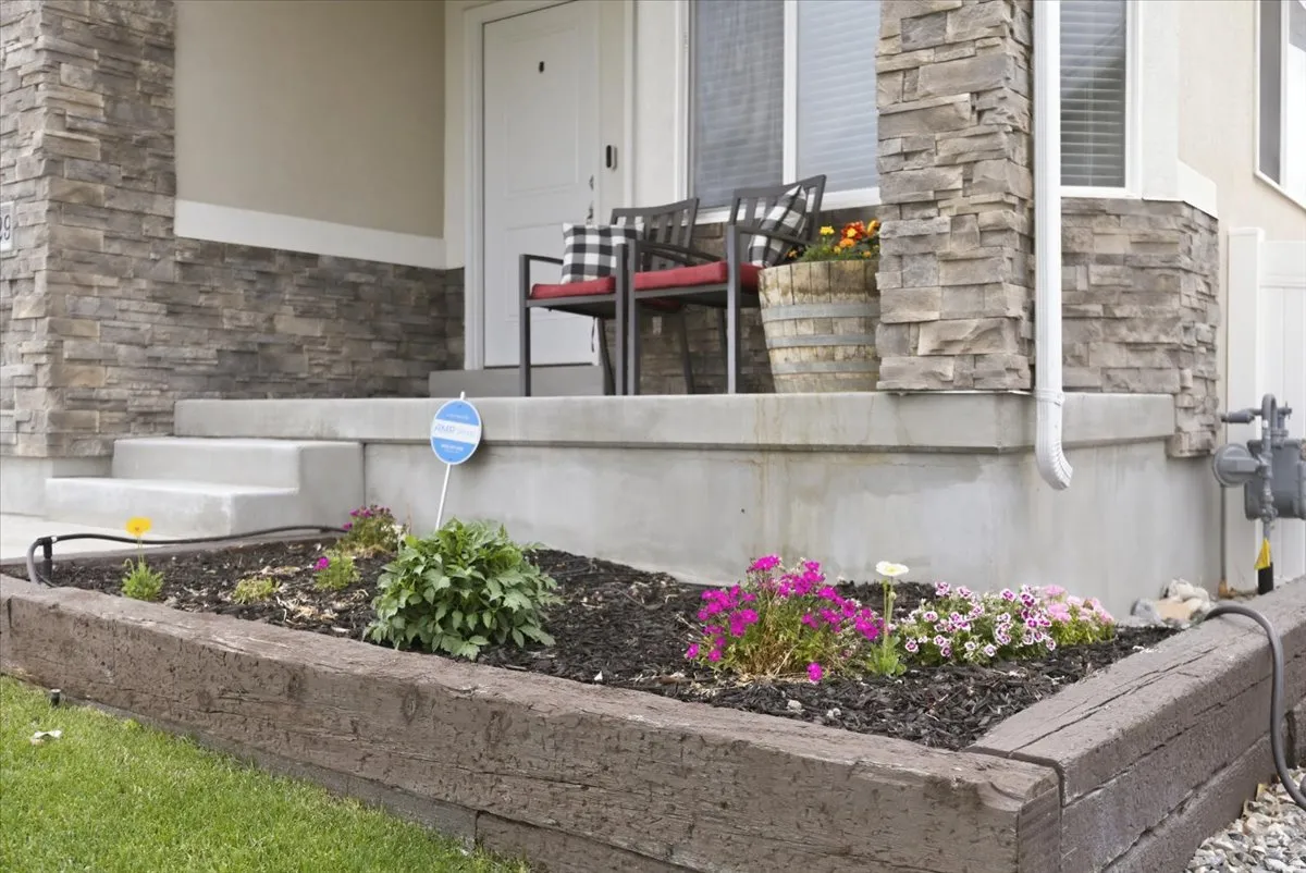 Stacked stone exterior accents, concrete steps, a raised flower bed with wood-finish landscaping timbers, and a white entry door