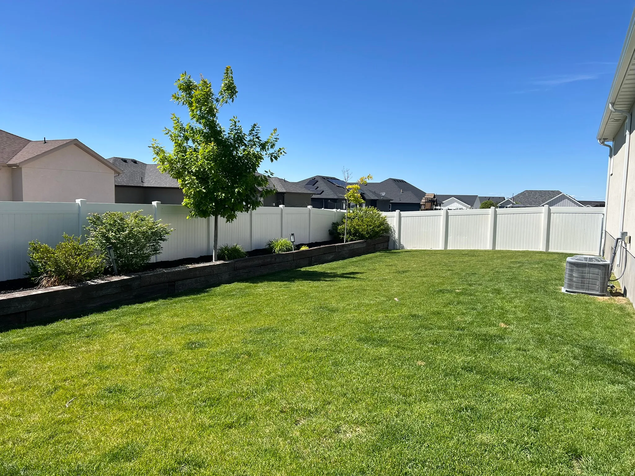 Expansive backyard featuring a lush lawn, white vinyl fencing, and a terraced planter with mature shrubs and a young tree