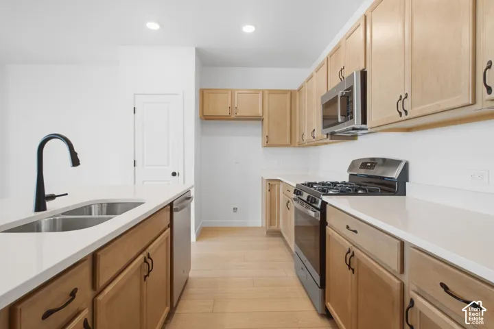 Kitchen featuring light-tone wood cabinetry, solid-surface countertops, stainless steel appliances, a center island with an undermount sink and matte black faucet, and wood-finish flooring