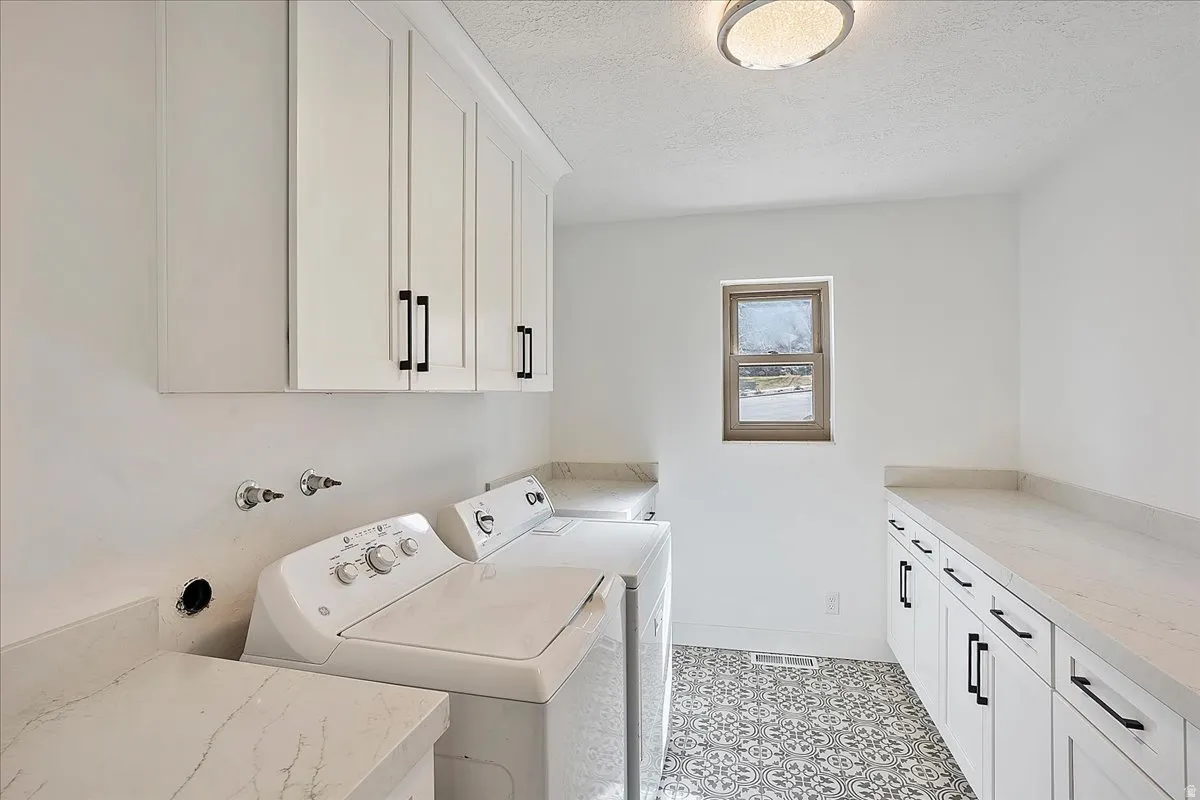 Bright laundry room featuring white cabinetry with matte black hardware, stone-finish countertops, and patterned floor tiling