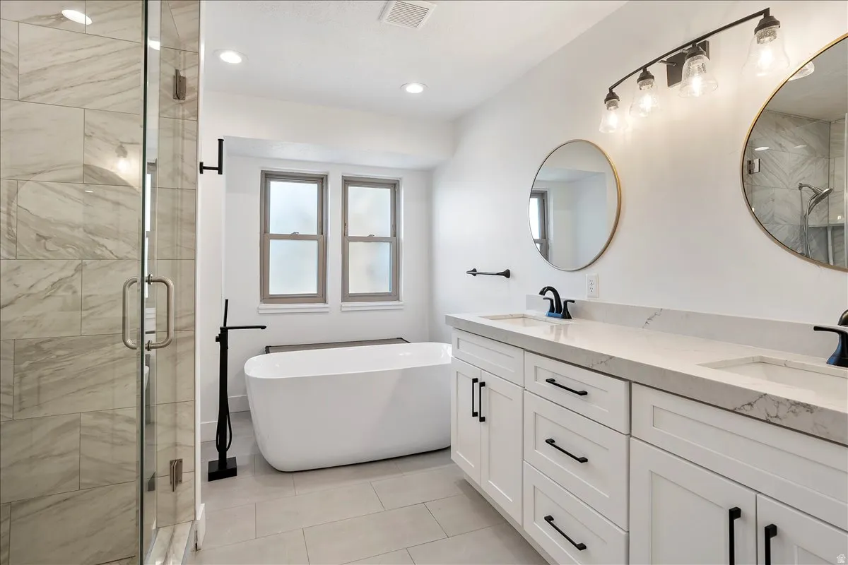 Contemporary bathroom featuring a freestanding soaking tub, a frameless glass shower enclosure with large format tile, a dual vanity with white cabinetry and stone countertops, and two round mirrors with brass-finish frames