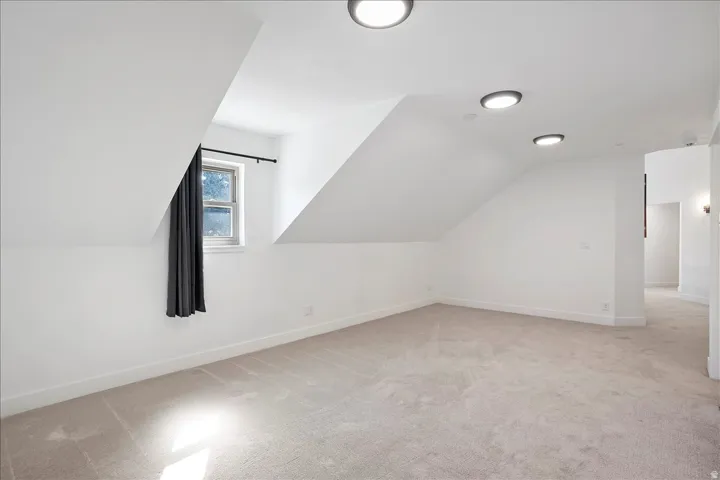 Spacious room featuring sloped ceilings, recessed lighting, neutral carpeting, white baseboards, and a window with a dark curtain