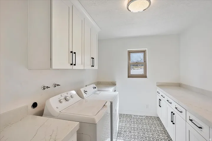 Bright laundry room featuring white cabinetry with matte black hardware, stone-finish countertops, and patterned floor tiling