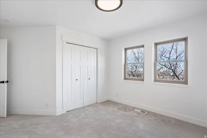 Bright room with light gray carpeting, two sash windows with gray frames, a white bi-fold closet, a white door with black hardware, and a flush-mount ceiling light