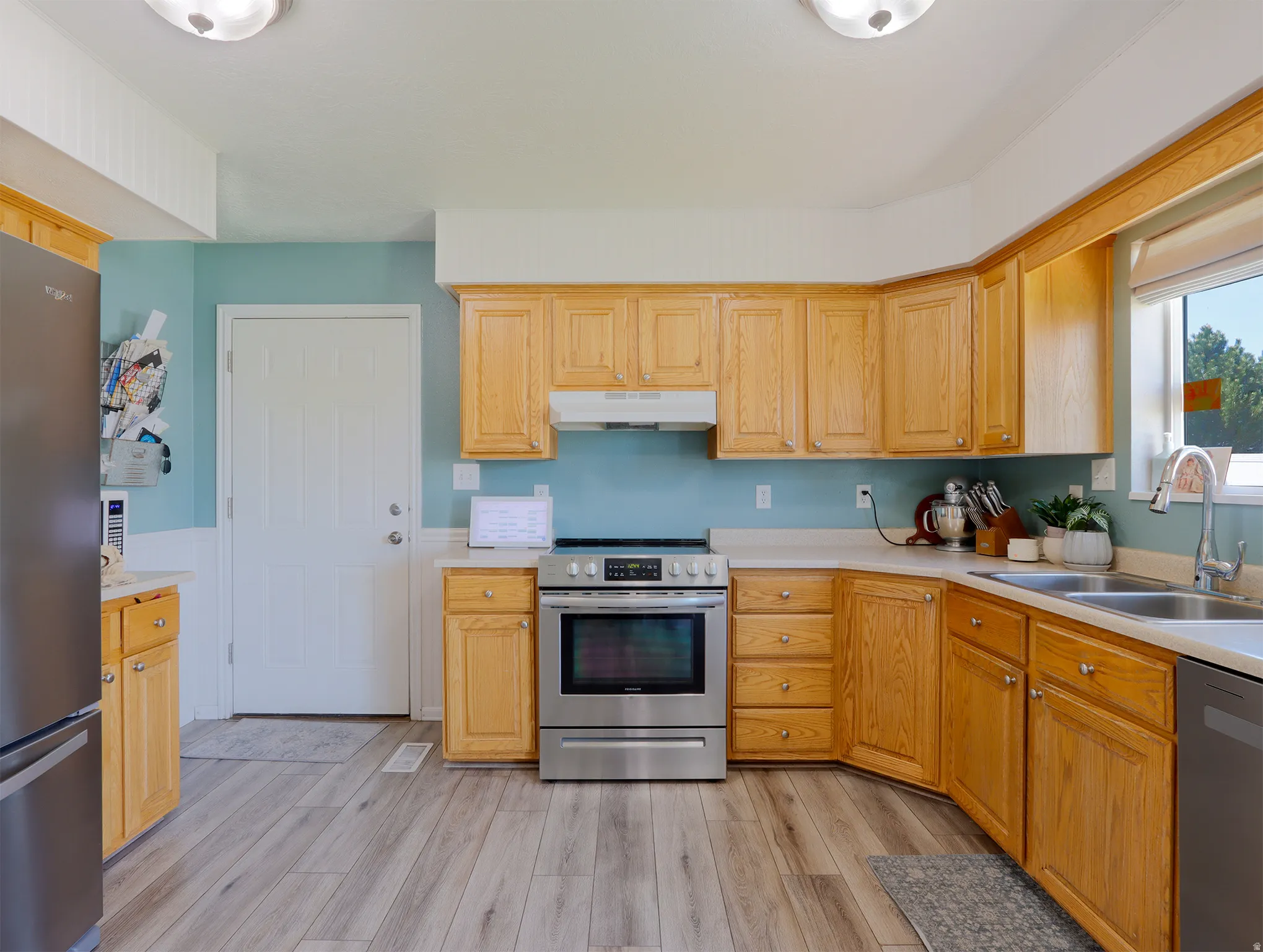 Kitchen featuring wood-finish flooring, light wood cabinetry, stainless steel appliances, and a window over the double basin sink