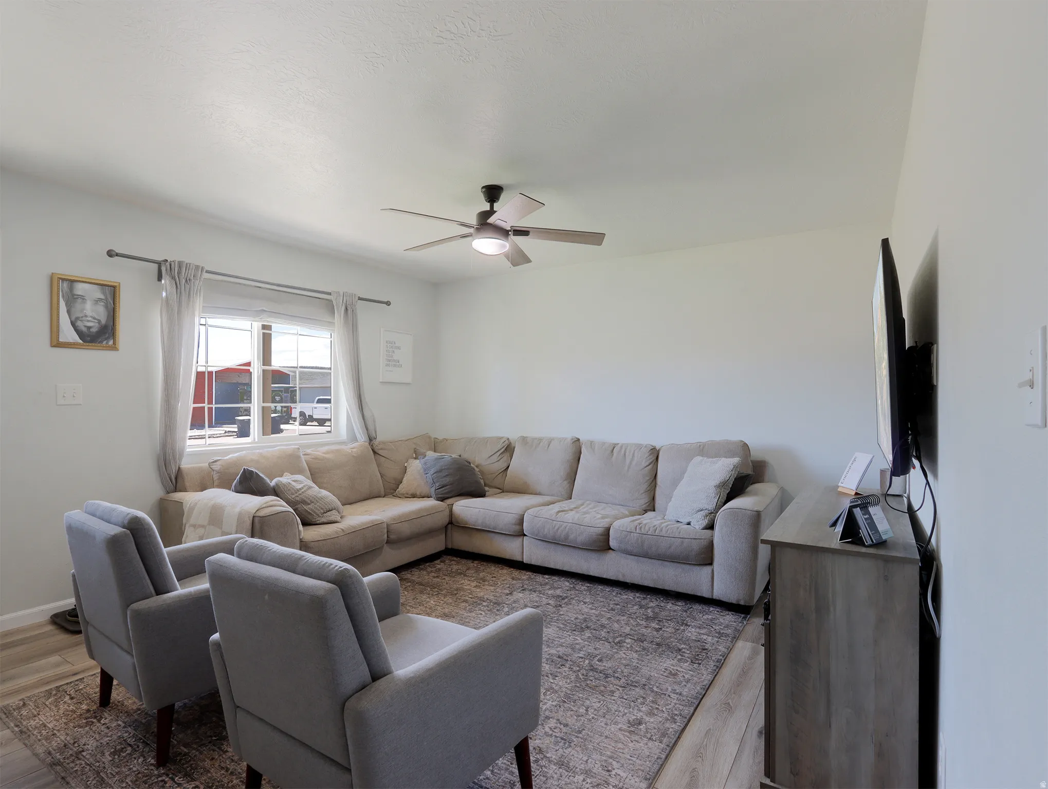 Bright living area featuring light-toned walls, wood-finish flooring, a ceiling fan with integrated lighting, and a large window providing natural illumination