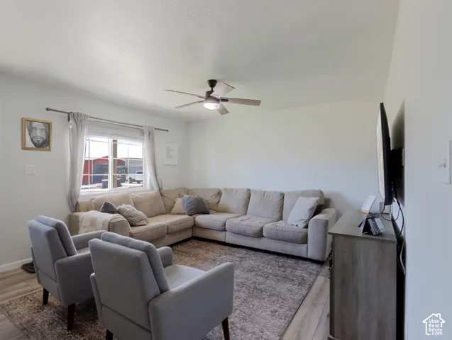Bright living area featuring light-toned walls, wood-finish flooring, a ceiling fan with integrated lighting, and a large window providing natural illumination