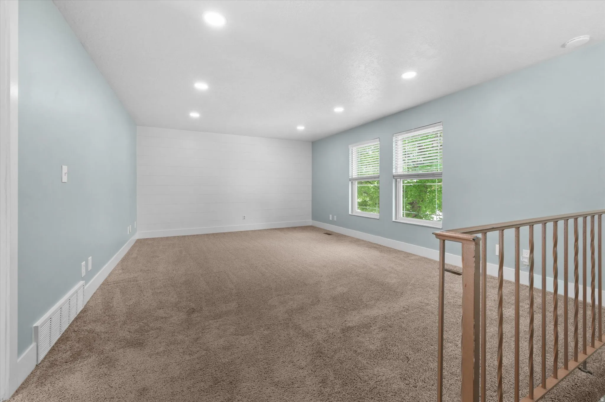 Spacious room featuring neutral carpeting, light blue painted walls, recessed lighting, and two windows with blinds