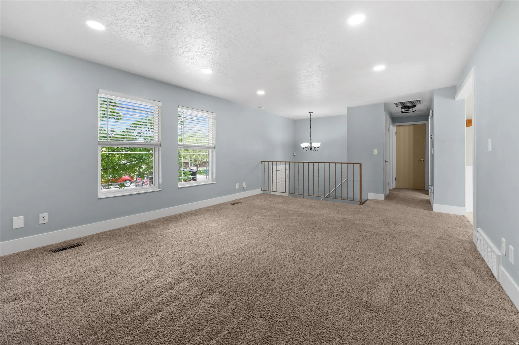 Spacious room with plush carpeting, two large windows with blinds, recessed lighting, and a partial baluster railing