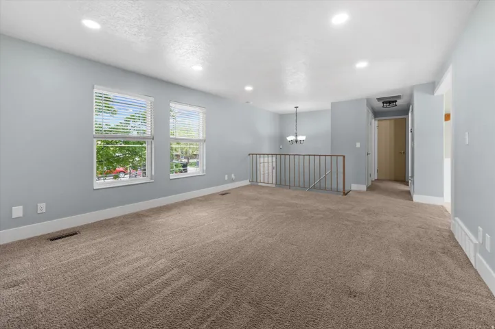 Spacious room with plush carpeting, two large windows with blinds, recessed lighting, and a partial baluster railing