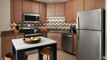 Contemporary kitchen featuring wood-finish cabinetry, stainless steel appliances, a chevron pattern backsplash, and stone countertops