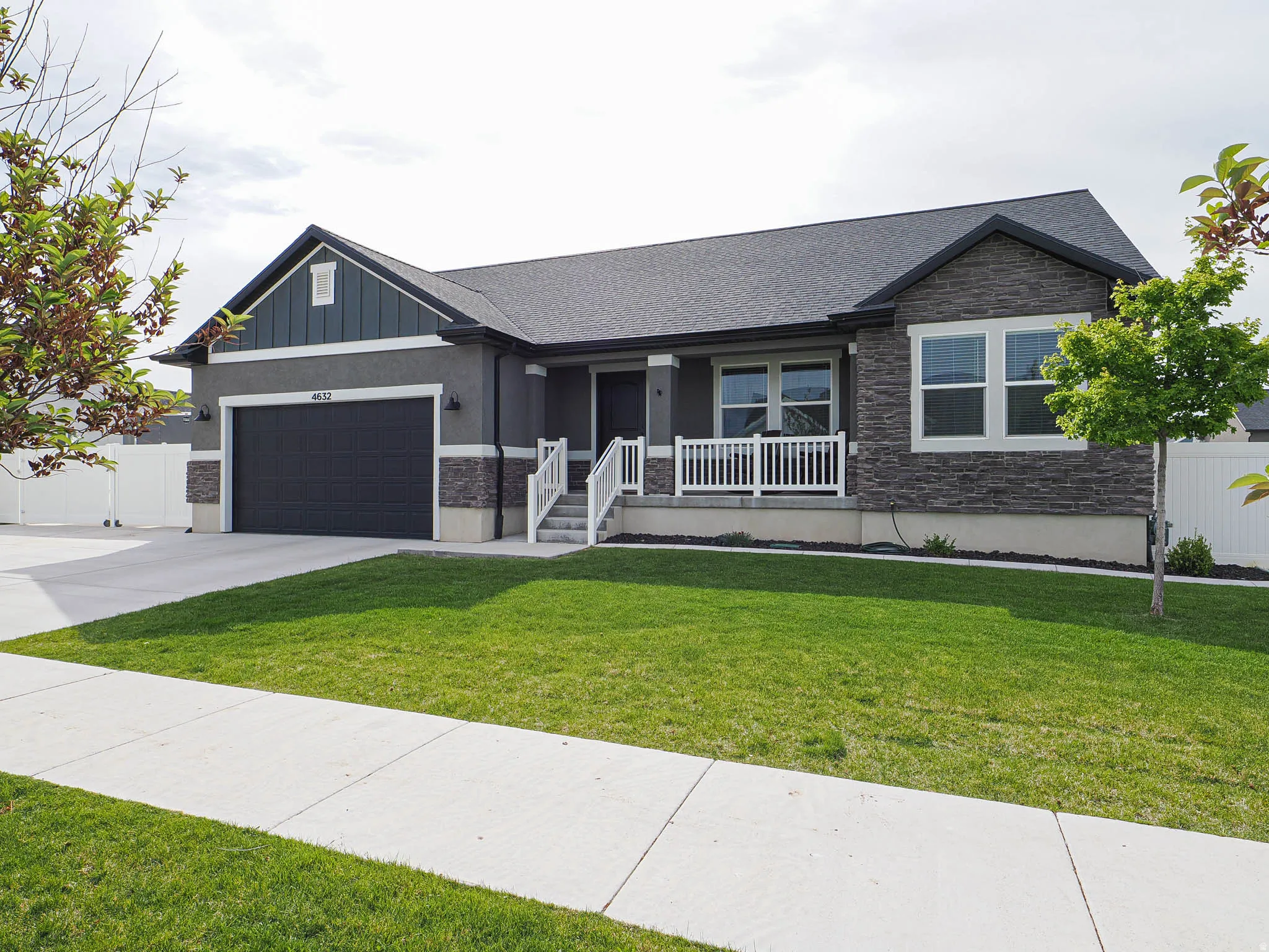 Contemporary ranch-style residence featuring a dark gray exterior, prominent stone accents, an attached two-car garage, white porch railings, and a meticulously maintained front lawn