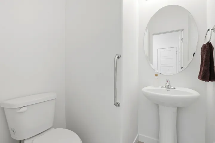 Bathroom right off kitchen pedestal sink with chrome fixtures and an oval mirror, complemented by a white toilet and a chrome grab bar