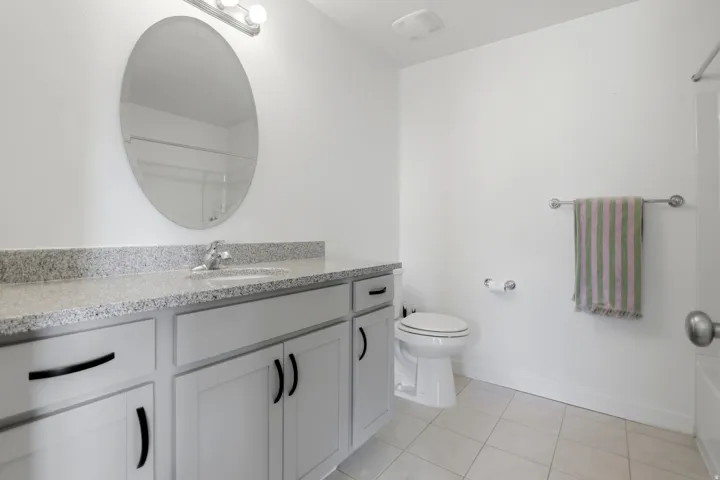 Bathroom featuring a vanity with a light-colored countertop, an oval mirror, white cabinetry with dark hardware, a toilet, and light-toned floor tiles