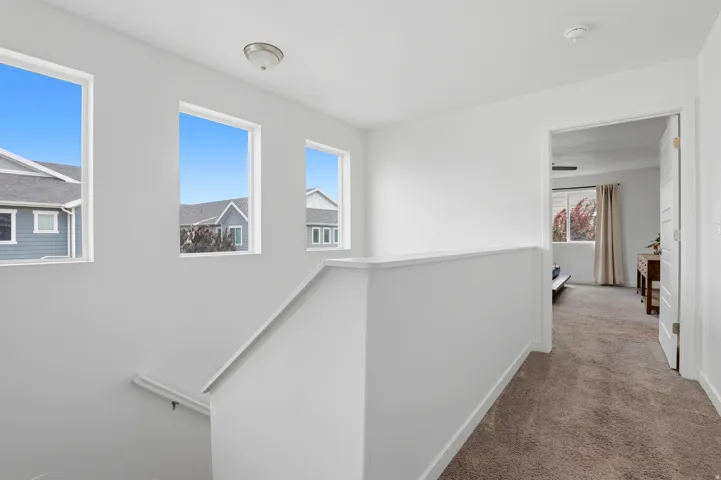 Upper-level hallway featuring a neutral color palette, three vertical windows, a ceiling-mounted light fixture, carpeted flooring, and a white interior door with panel detailing