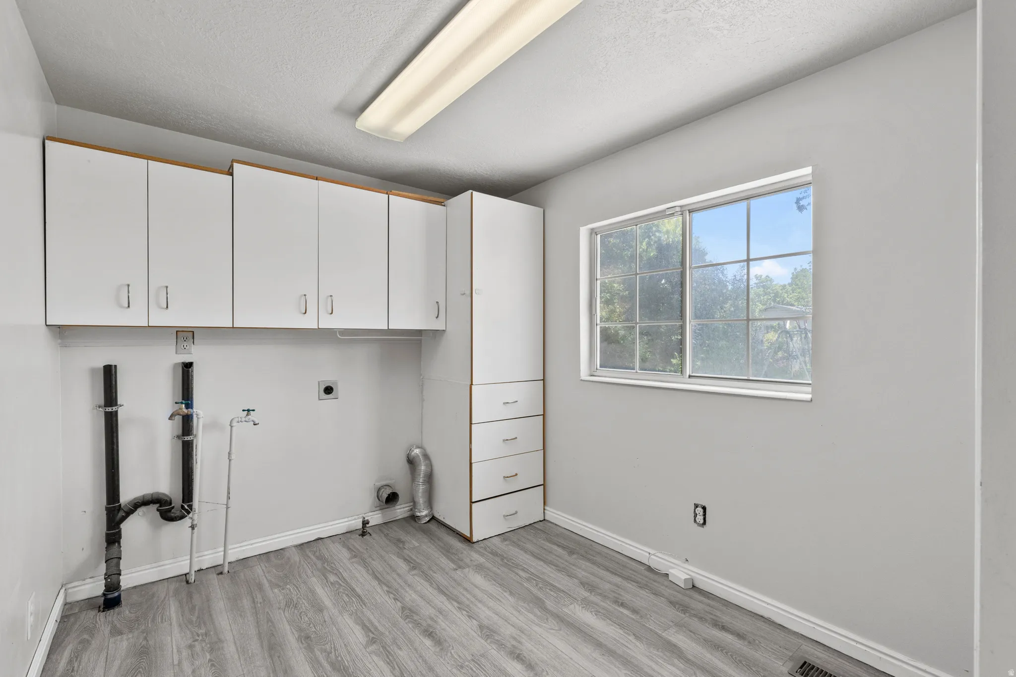 Laundry room featuring wood-finish flooring, overhead cabinetry, a built-in storage tower with drawers, and a large window. Closet with shelves also.  Gas and electric for dryer.  What great space; you could even have a craft room in here!
