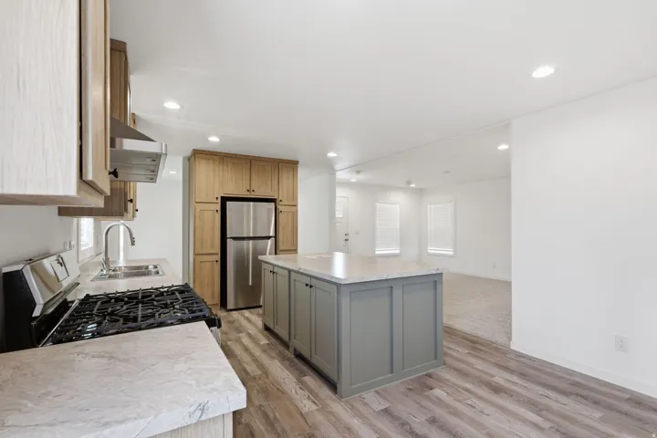 Kitchen featuring wood-finish flooring, a central island with gray cabinetry and light-toned countertop, stainless steel refrigerator, and a gas range with an overhead vent hood