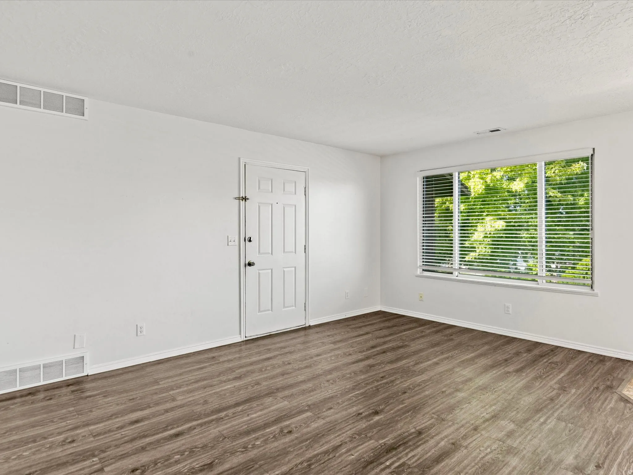 Spacious room with wood-finish flooring, white baseboards, and a large window featuring horizontal blinds