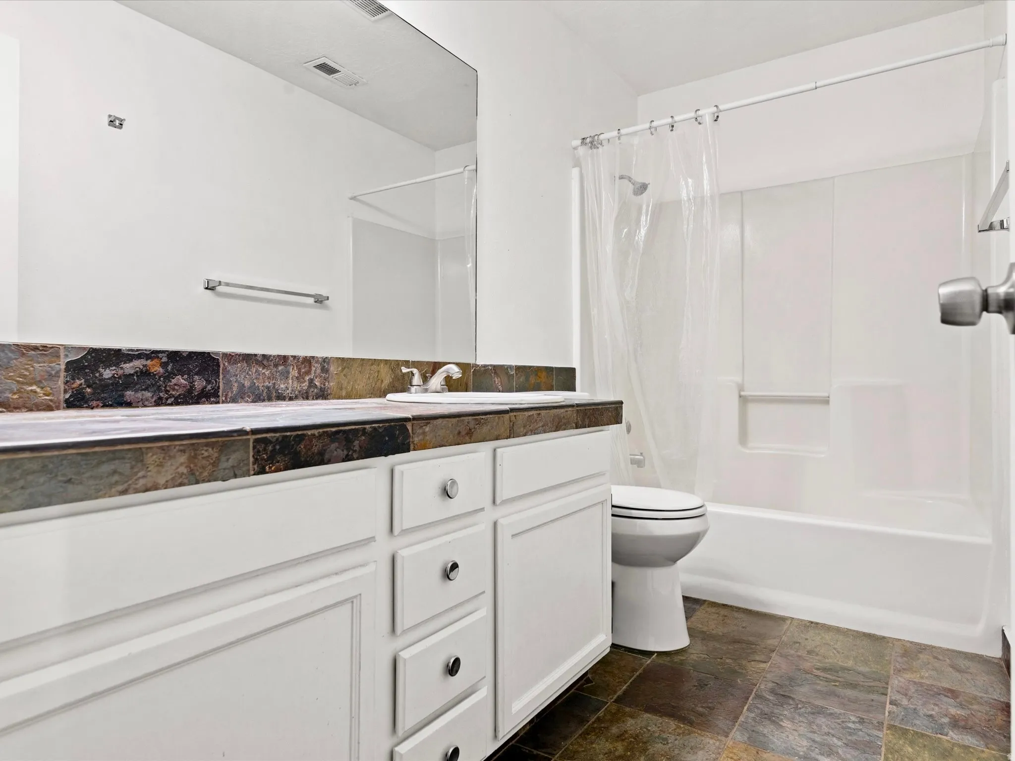 Bathroom featuring a white vanity with multiple drawers, a slate-tiled countertop and backsplash, an integrated sink, a large wall-mounted mirror, and a white bathtub with a shower and a clear shower curtain