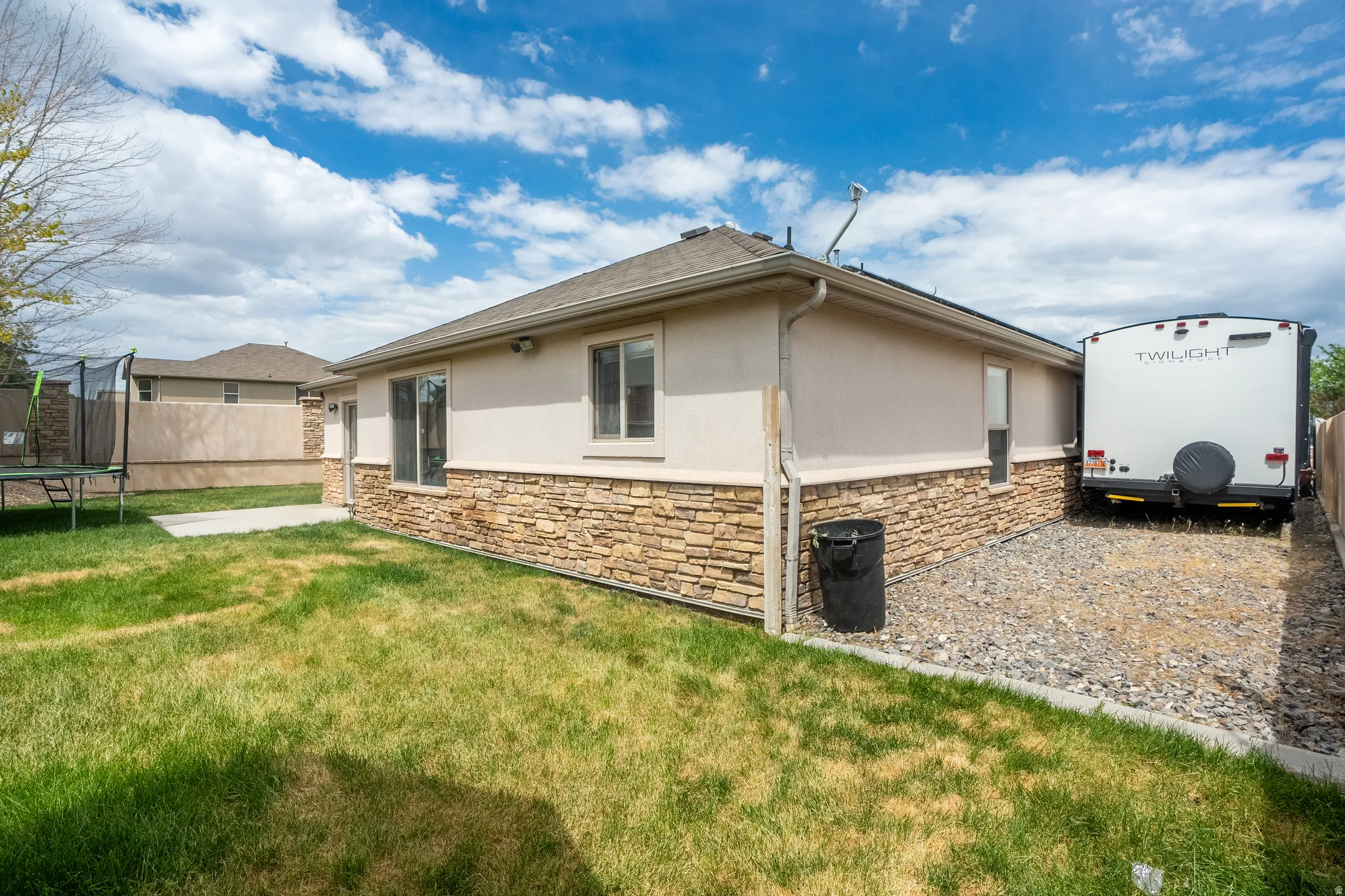 Rear exterior featuring stacked stone accents, stucco siding, a gravel-covered RV parking area, a concrete patio, and a grass lawn