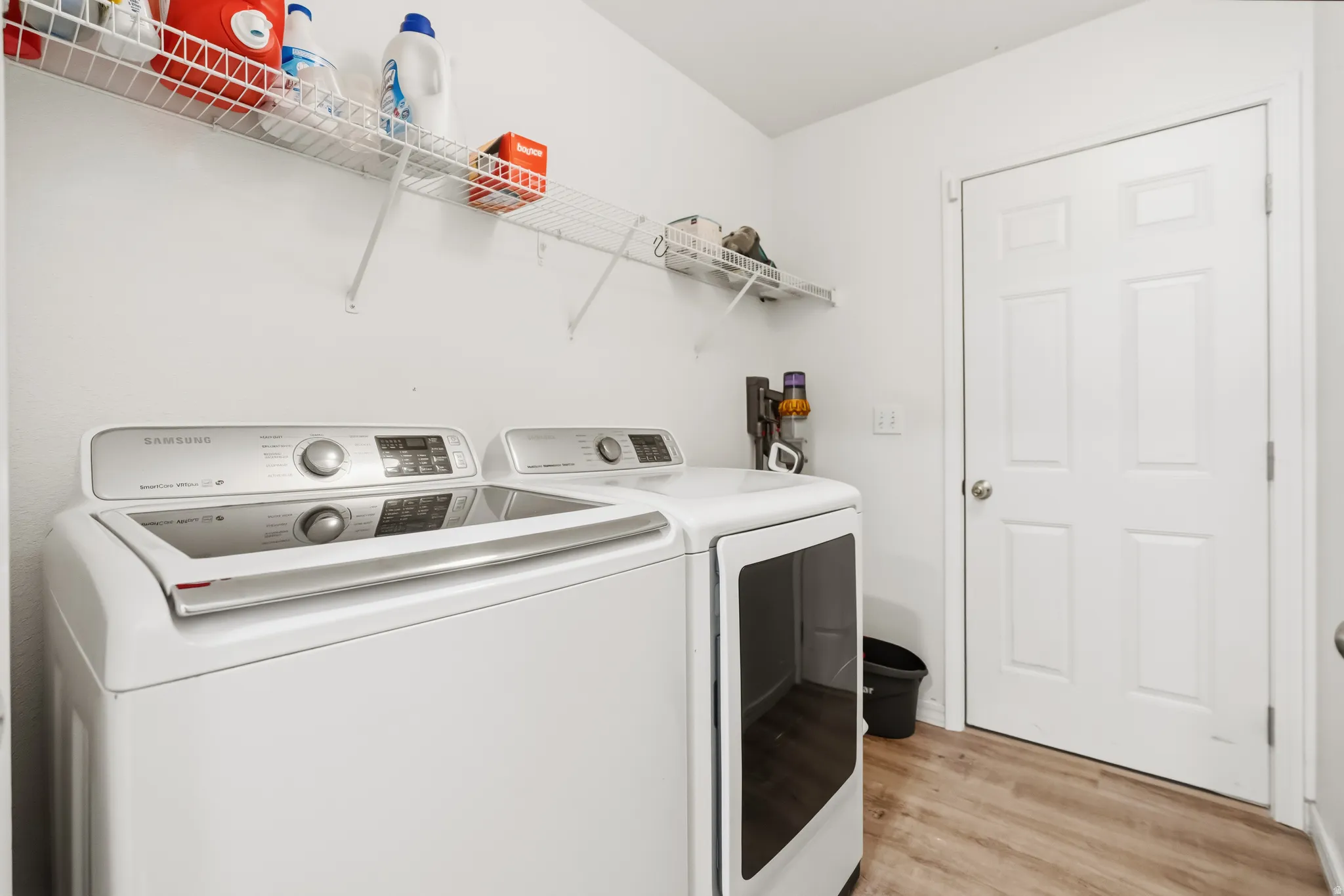 Dedicated laundry area featuring a white top-load washing machine and front-load dryer set, wire shelving, white walls, and wood-finish flooring