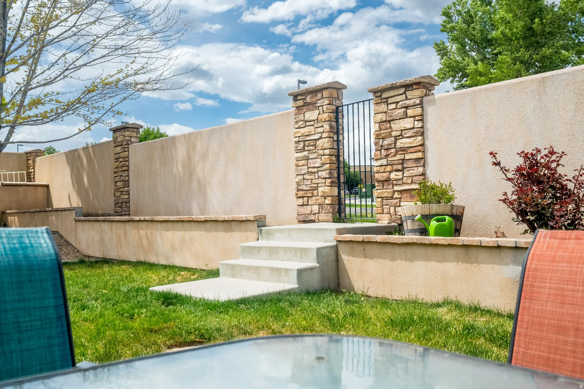 Manicured lawn area featuring a stucco privacy wall with stone-clad pillars, an ornamental iron gate, and tiered concrete steps