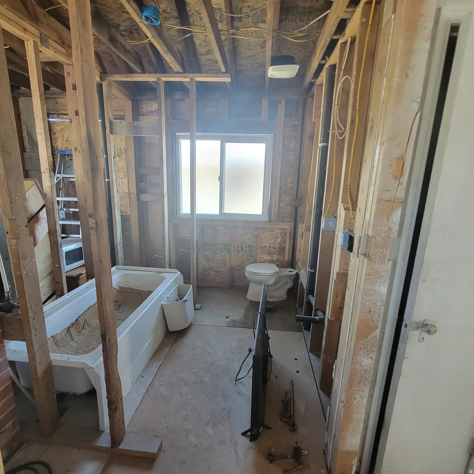Bathroom space undergoing renovation, featuring exposed wooden framing, an existing bathtub, and a window with frosted panes
