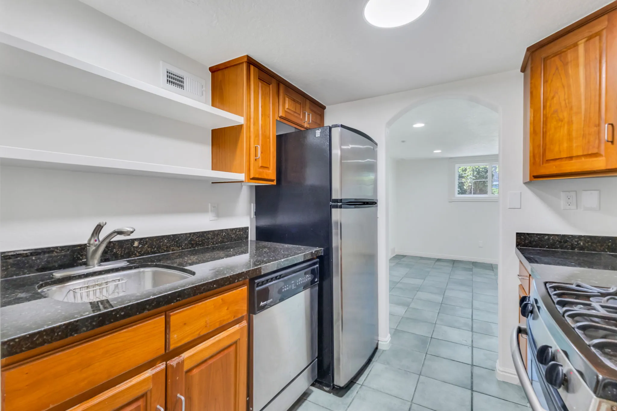 Kitchen featuring a stainless steel sink, dark granite-style countertops, wood cabinetry, and open shelving