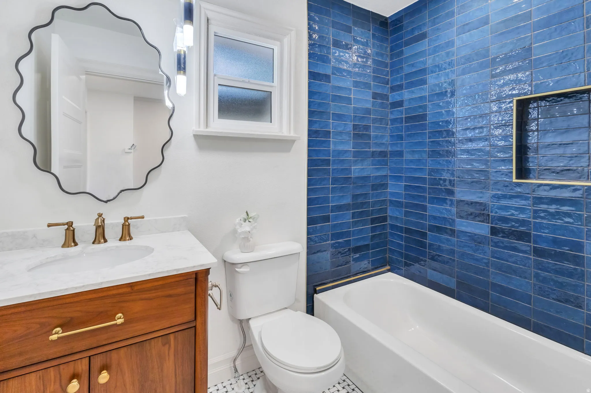 Bathroom featuring a white bathtub with a blue tiled surround, built-in shelving niche, wood-finish vanity with a white countertop, and brass-finish fixtures