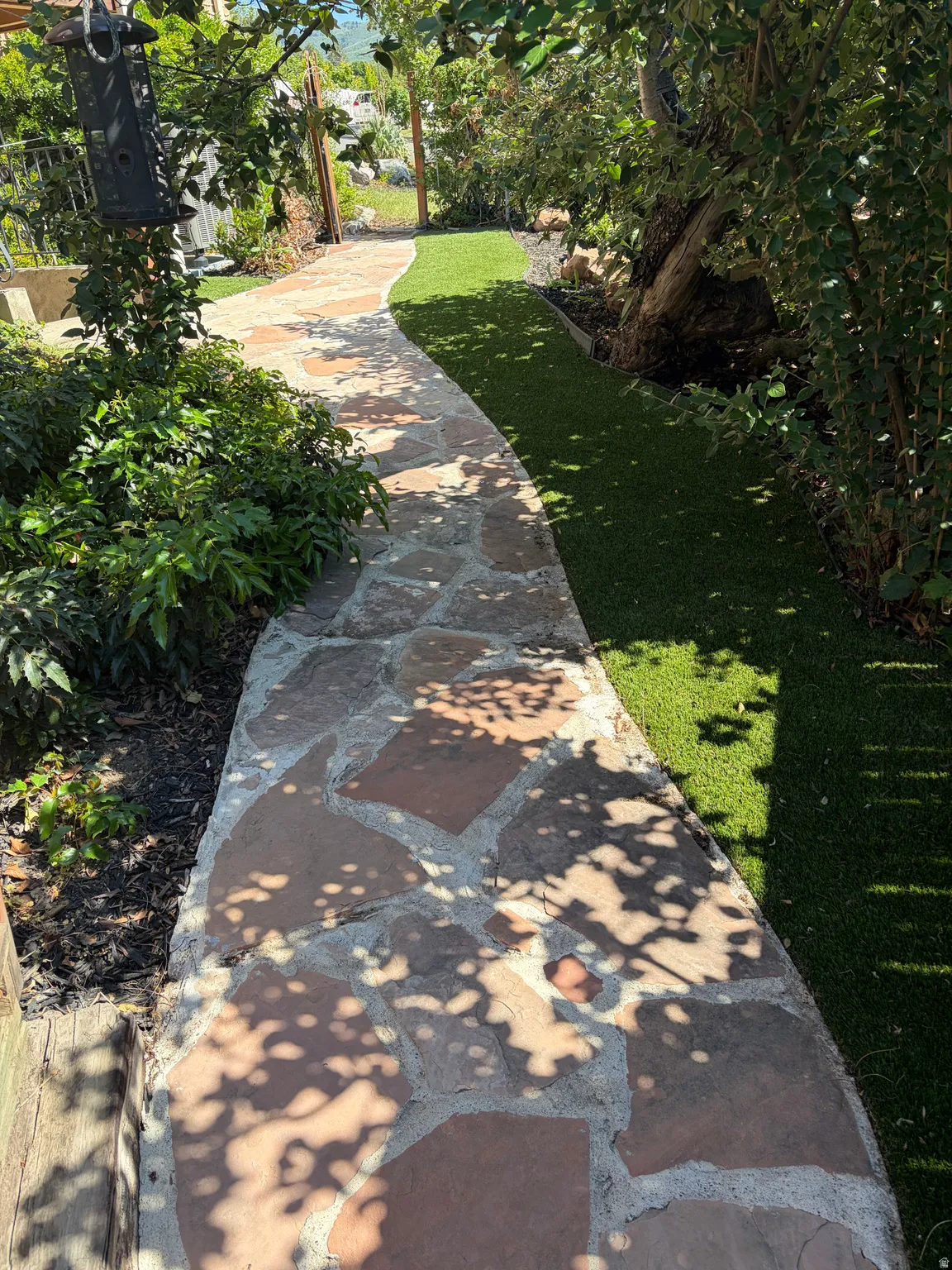 Curved flagstone pathway bordered by dense green foliage and a strip of artificial turf