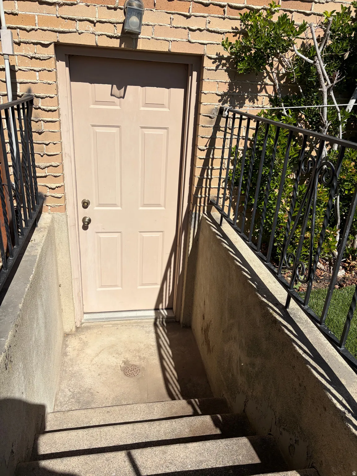 Exterior entry featuring a tan paneled door, brick siding, wrought iron railings, concrete stairs, and an overhead light fixture