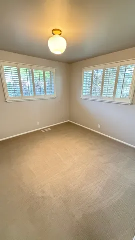 Spacious room featuring neutral carpeting, two windows with plantation shutters, light beige wall paint, white baseboards, and a central ceiling light fixture