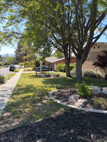 Front yard featuring mature trees, landscaped planting beds with rocks, a sidewalk, and a brick exterior home with an attached garage