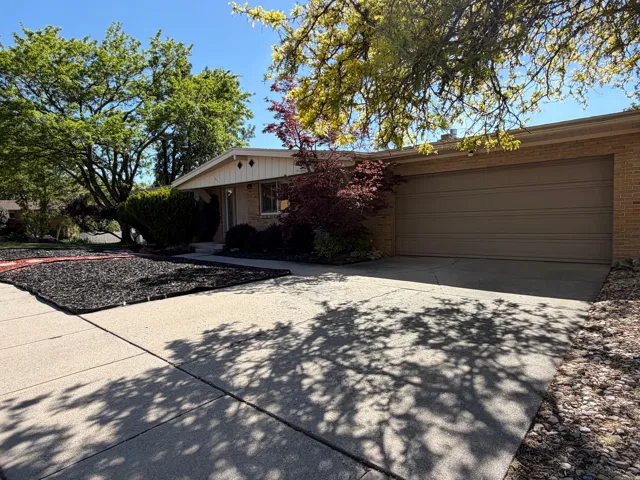 Attached two-car garage with a concrete driveway, brick facade, mature landscaping, and decorative bark ground cover