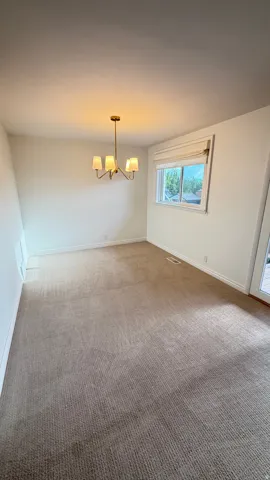 Open concept room featuring a modern brass chandelier with four frosted glass shades, a large window with a Roman shade, and light brown carpeting