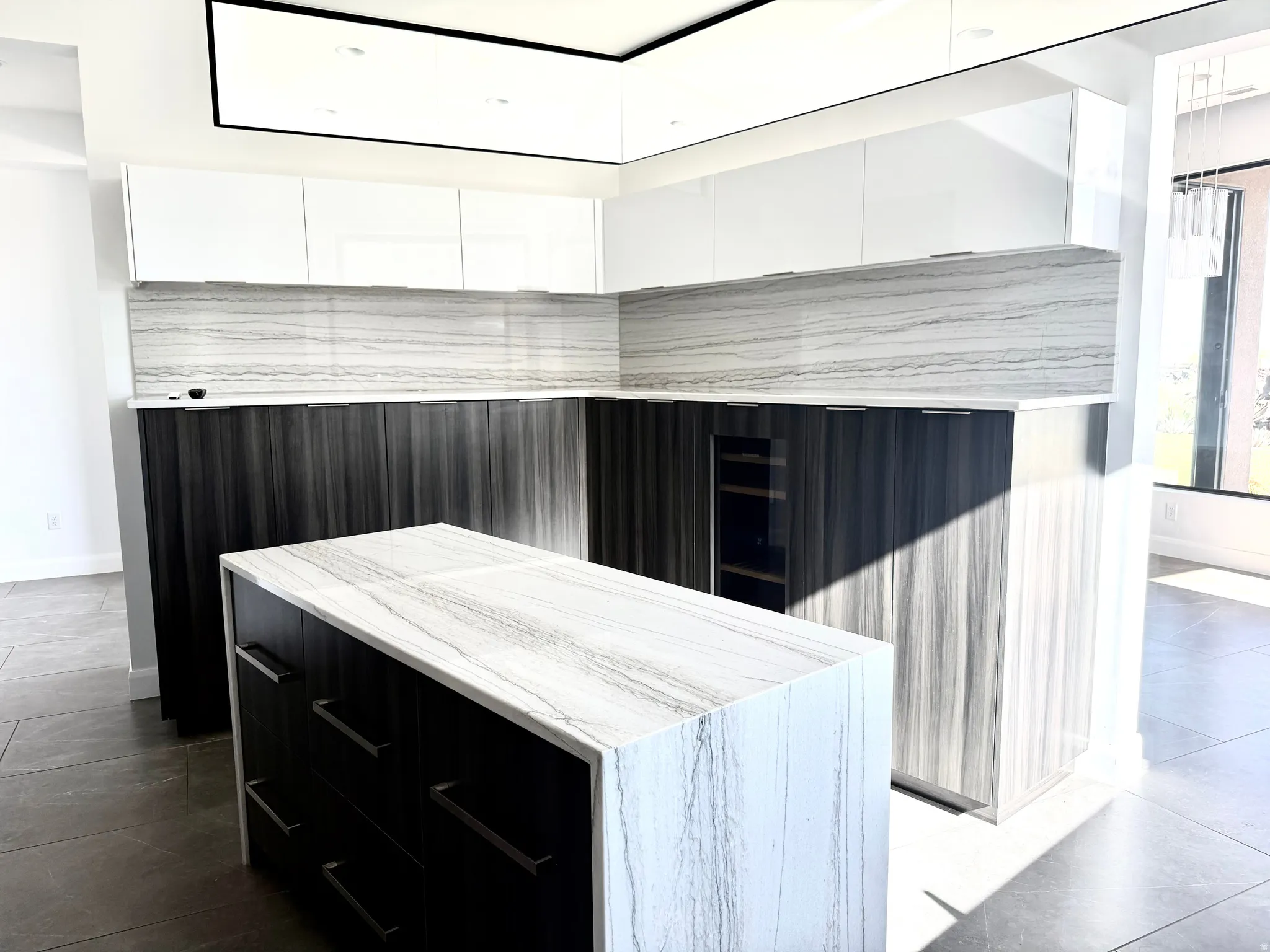 Contemporary kitchen featuring an island with a waterfall-edge countertop, two-tone cabinetry with white upper and dark wood-finish lower units, full-height stone slab backsplash, and a built-in beverage cooler