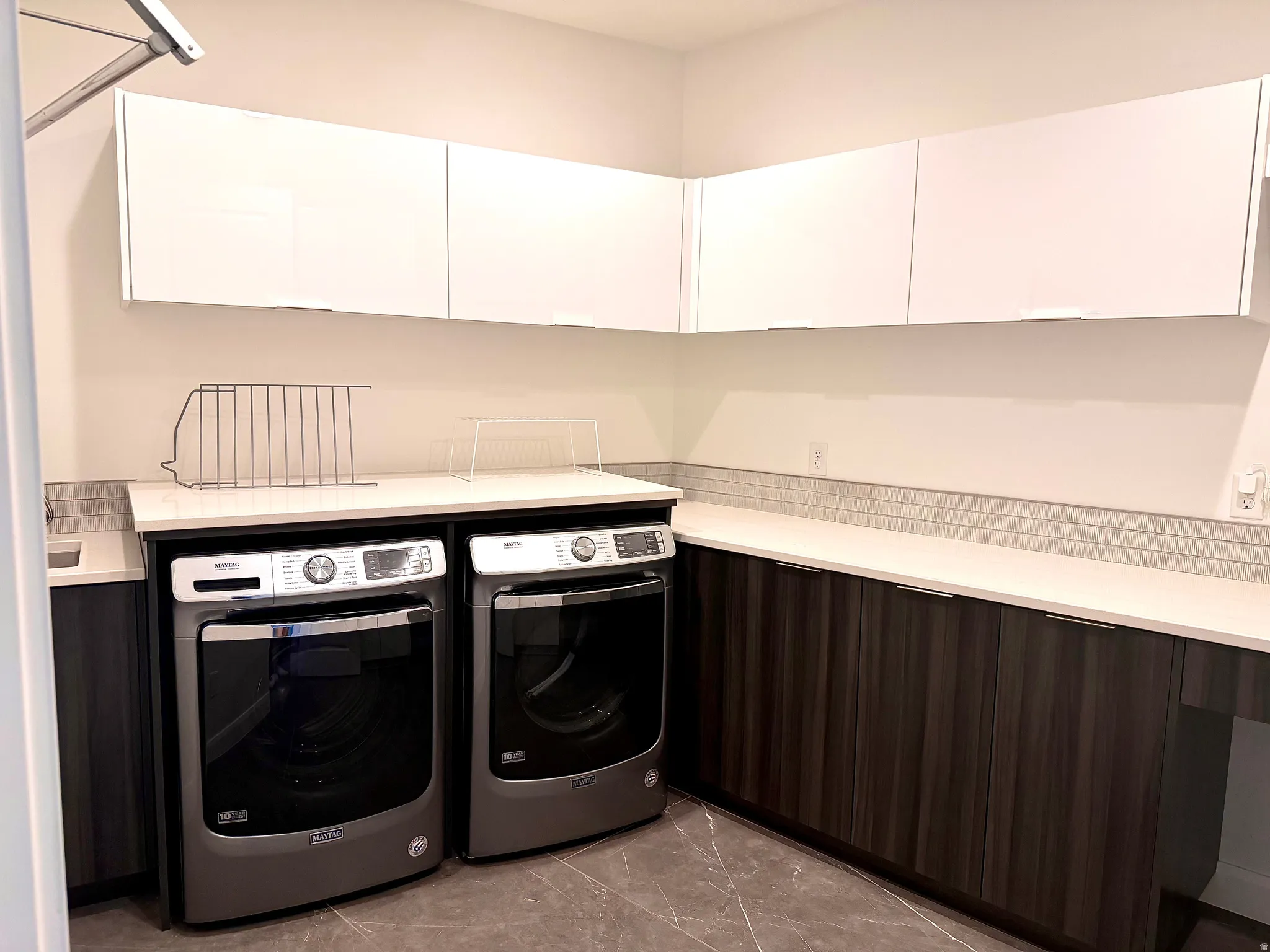 Laundry area featuring white upper cabinetry, dark wood-finish lower cabinetry, white countertops, and a gray tile backsplash