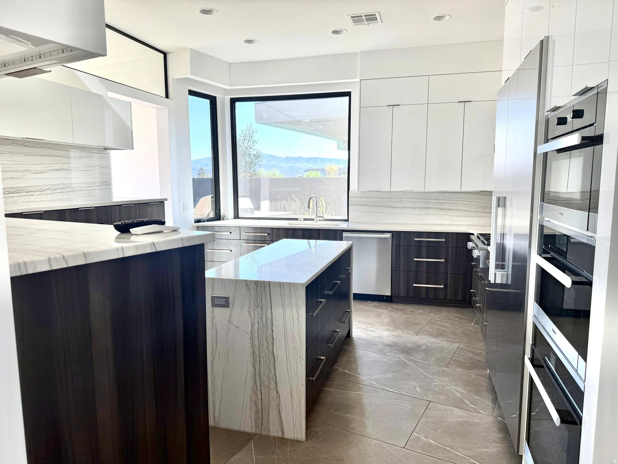 Contemporary kitchen featuring a waterfall island with stone countertop, dark wood-finish cabinetry, integrated stainless steel appliances, and large windows