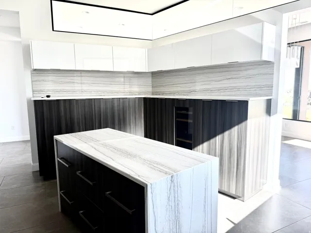Contemporary kitchen featuring an island with a waterfall-edge countertop, two-tone cabinetry with white upper and dark wood-finish lower units, full-height stone slab backsplash, and a built-in beverage cooler