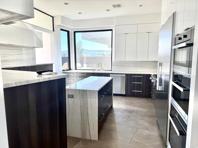 Contemporary kitchen featuring a waterfall island with stone countertop, dark wood-finish cabinetry, integrated stainless steel appliances, and large windows